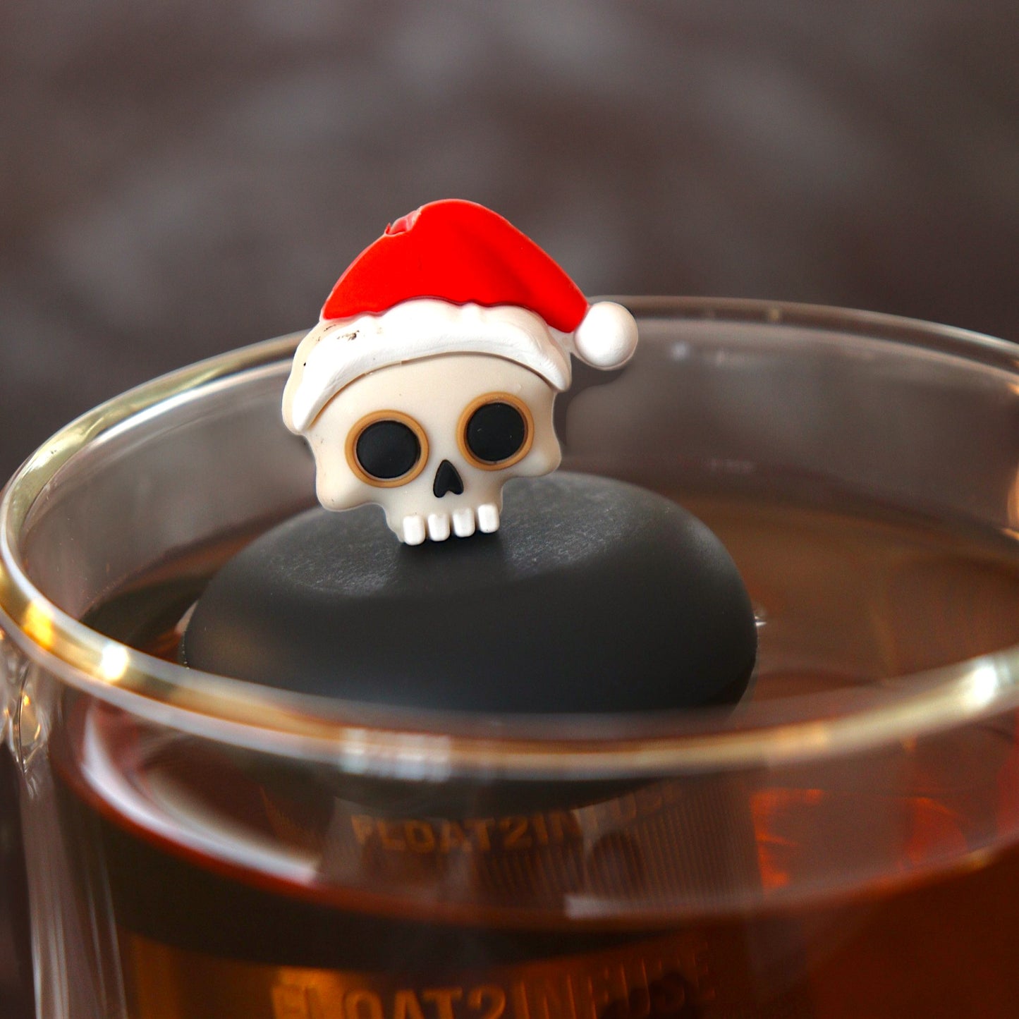 skull with a santa hat tea infuser for loose leaf tea with a fine mesh stainless steel tea infuser that reads FLOAT2INFUSE on a black base  floating in a glass tea mug