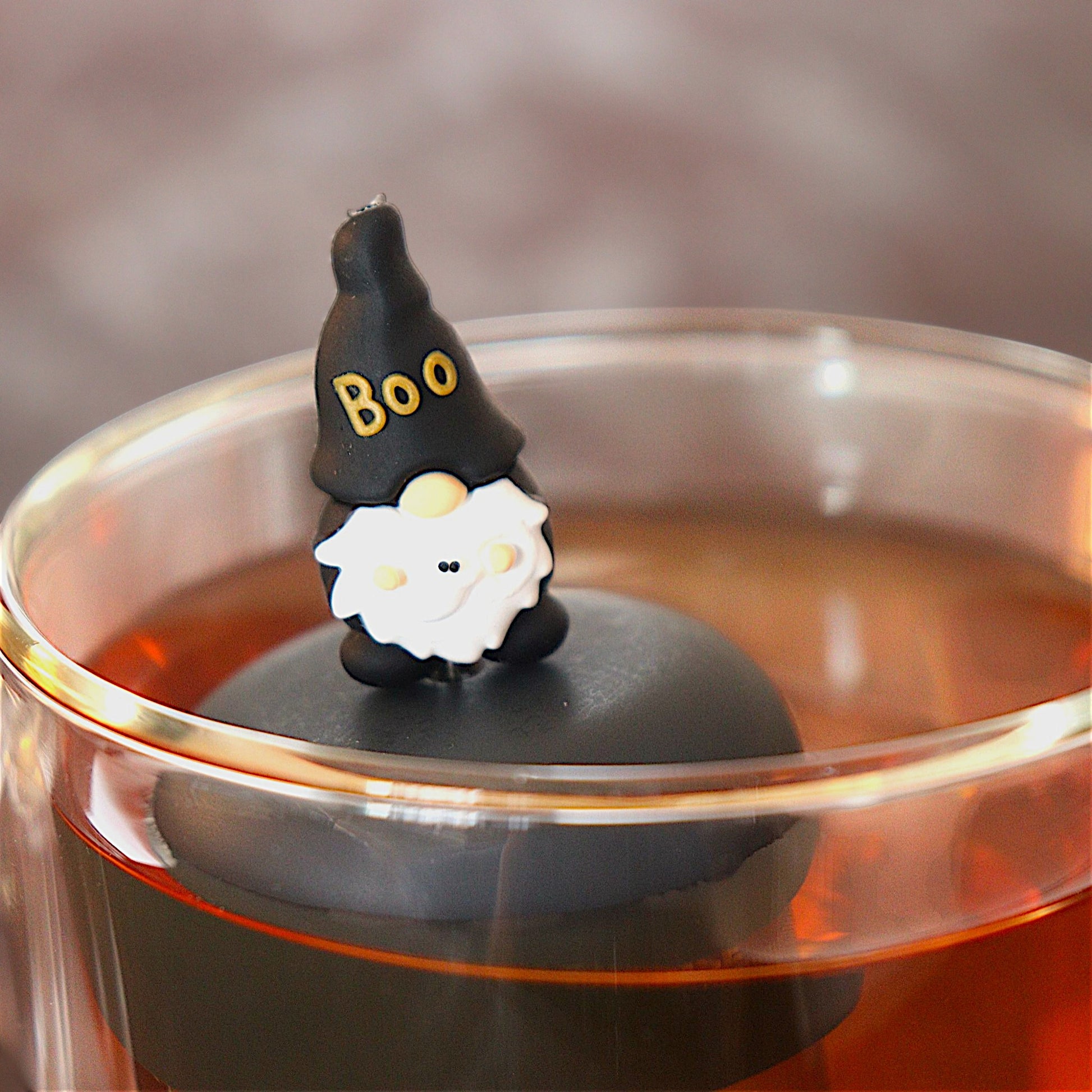 black gnome with a hat that reads "boo" holding a ghost tea infuser for loose leaf tea  with a fine mesh stainless steel tea infuser that reads FLOAT2INFUSE on a black base floating in a glass tea mug
