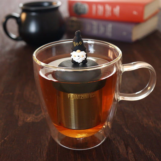 black gnome with a hat that reads "boo" holding a ghost tea infuser for loose leaf tea  with a fine mesh stainless steel tea infuser that reads FLOAT2INFUSE on a black base floating in a glass tea mug with a black cauldron mug and books in the background