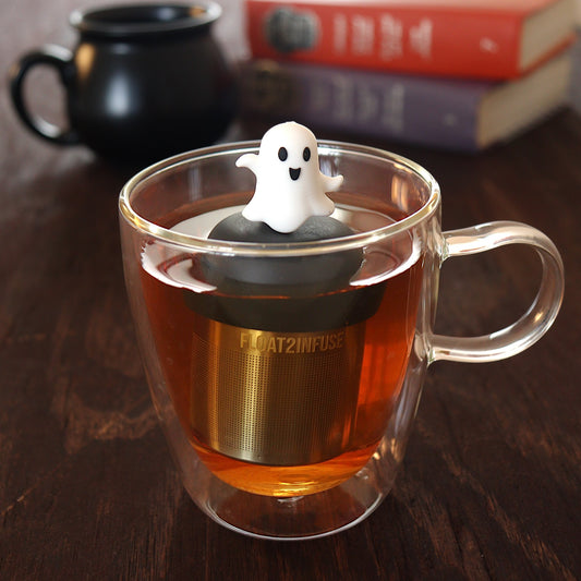 dancing white ghost tea infuser for loose leaf tea with a fine mesh stainless steel tea infuser that reads FLOAT2INFUSE on a black base  floating in a glass tea mug with a black cauldron mug and books in the background