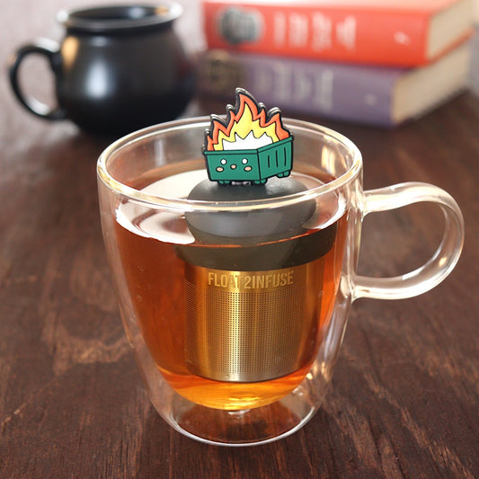 flames erupting out of the center of a happy green dumpster tea infuser for loose leaf tea with a fine mesh stainless steel tea infuser that reads FLOAT2INFUSE on a black base  floating in a glass tea mug with a black cauldron mug and books in the background