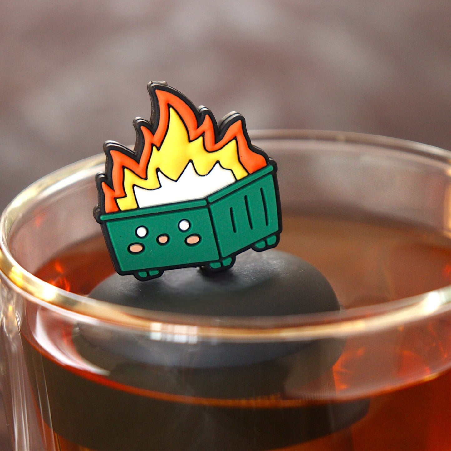 flames erupting out of the center of a happy green dumpster tea infuser for loose leaf tea with a fine mesh stainless steel tea infuser that reads FLOAT2INFUSE on a black base floating in a glass tea mug