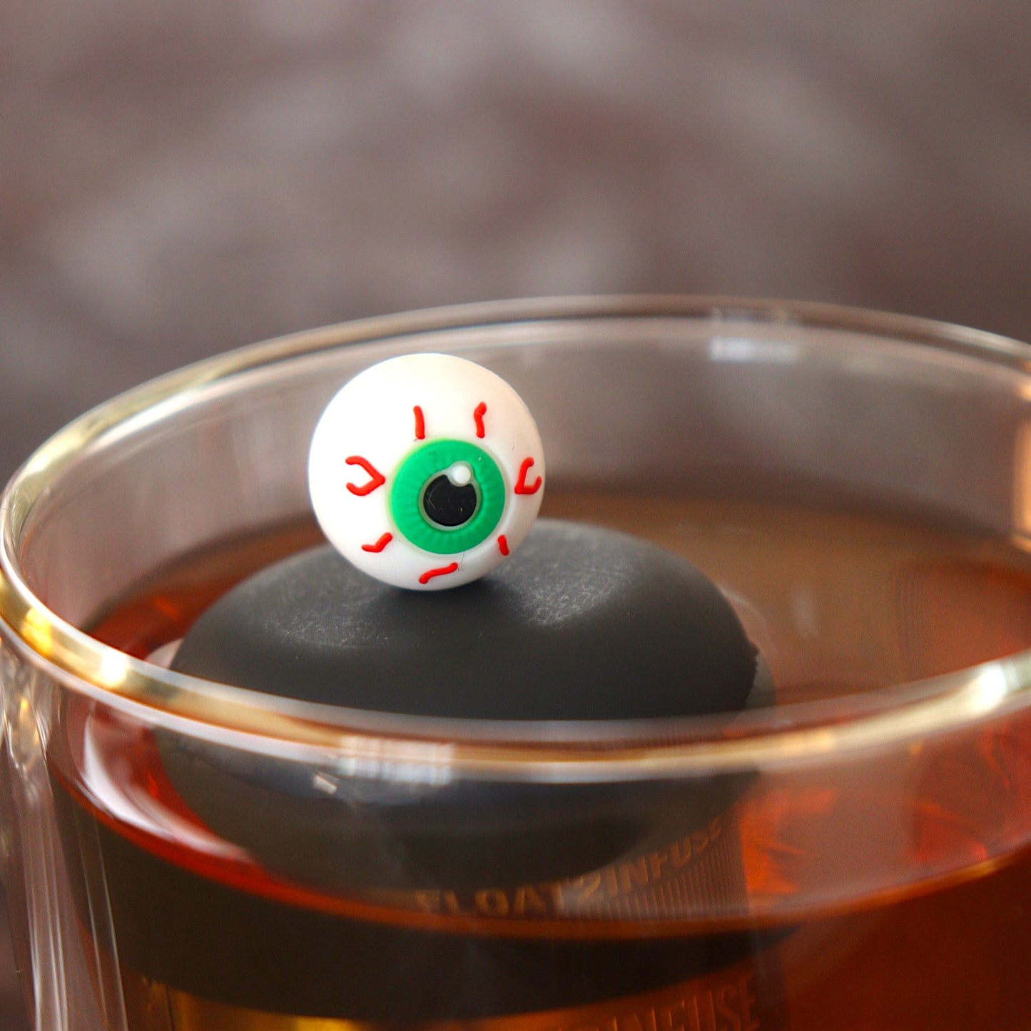green eyed eyeball tea infuser for loose leaf tea  with a fine mesh stainless steel tea infuser that reads FLOAT2INFUSE on a black base floating in a glass tea mug
