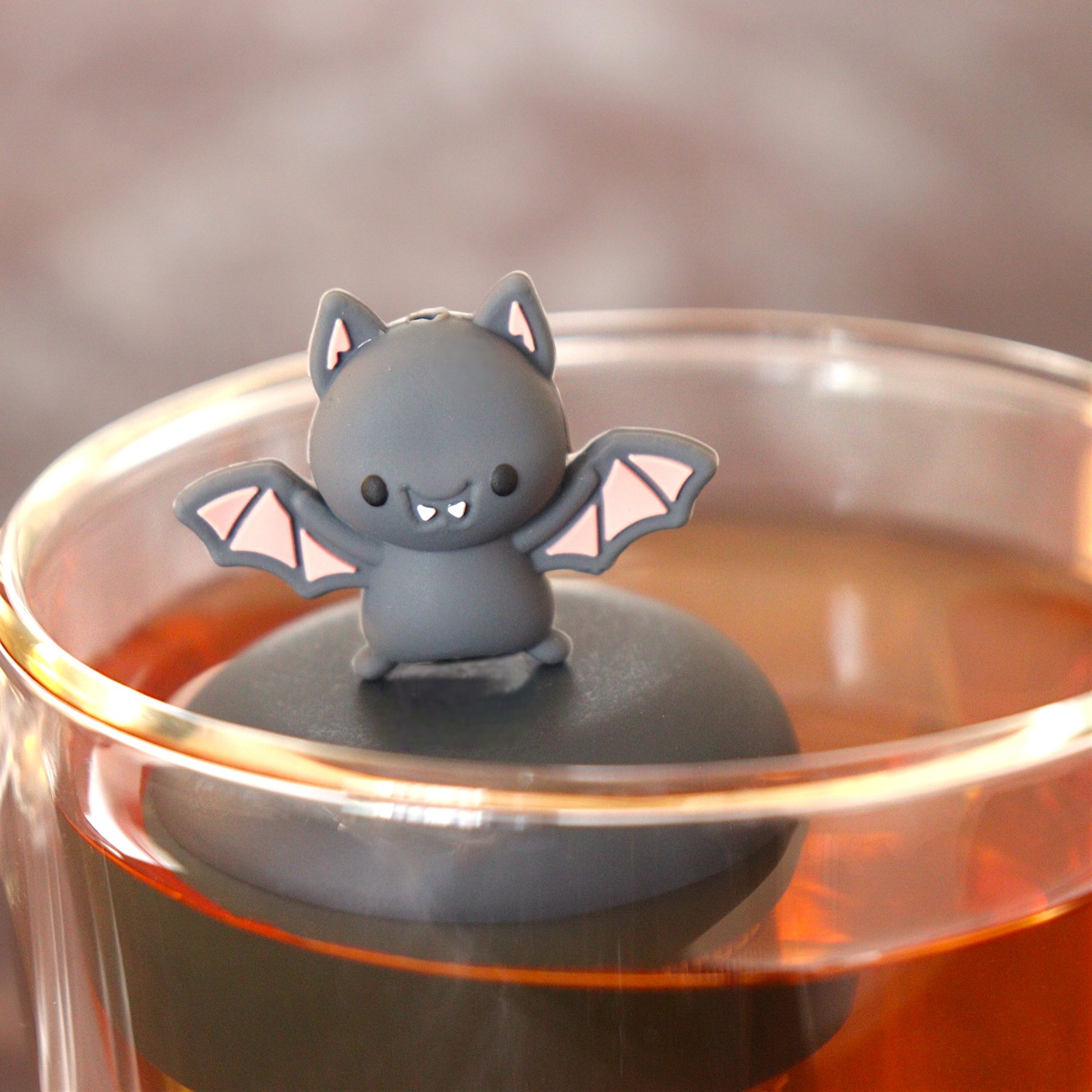 smiling grey and pink bat animal tea infuser for loose leaf tea  with a fine mesh stainless steel tea infuser that reads FLOAT2INFUSE on a black base floating in a glass tea mug