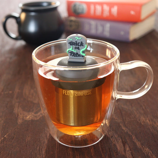 green vines wrap around a halloween themed tombstone with text that reads "trick or treat" tea infuser for loose leaf tea with a fine mesh stainless steel tea infuser that reads FLOAT2INFUSE on a black base  floating in a glass tea mug with a black cauldron mug and books in the background