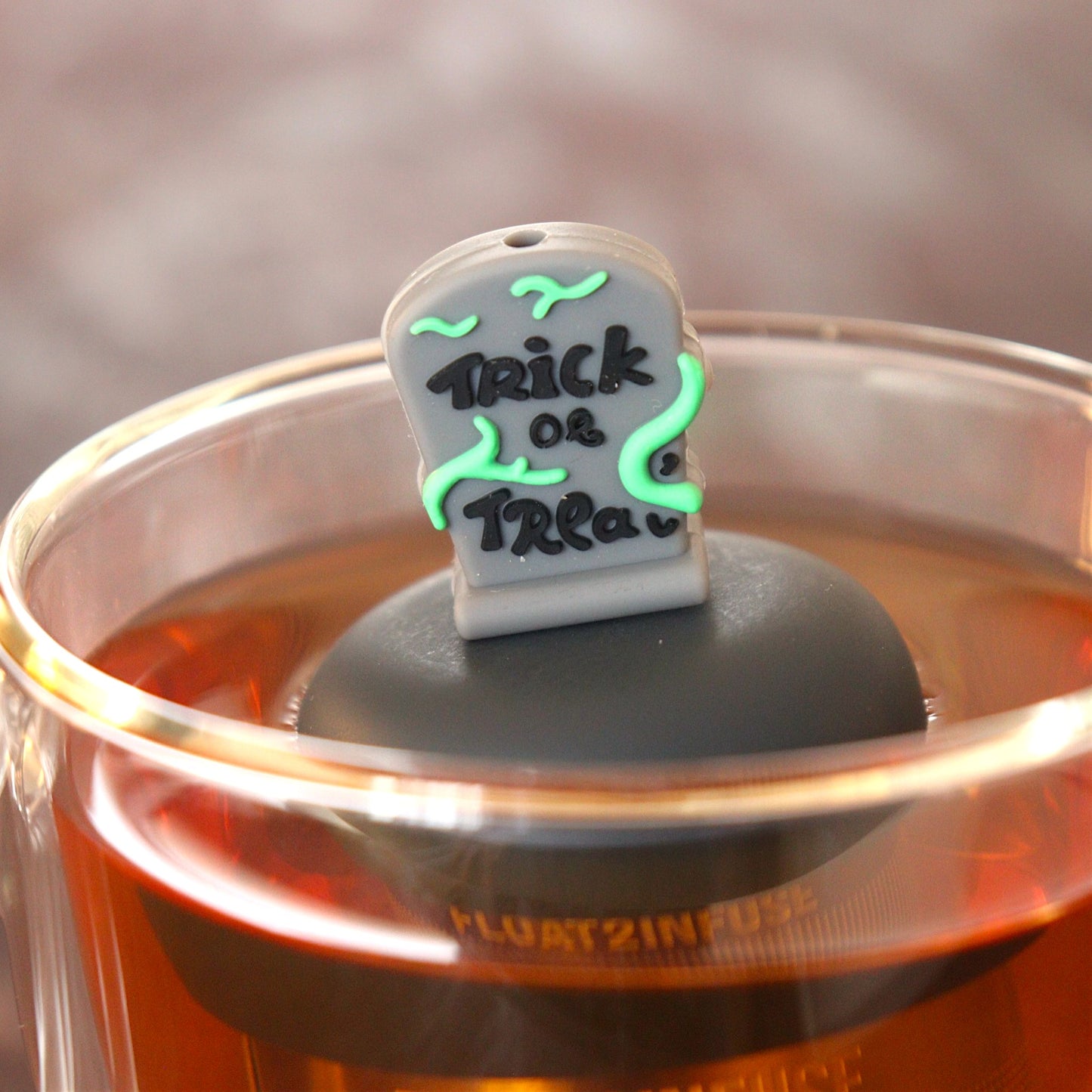 green vines wrap around a halloween themed tombstone with text that reads "trick or treat" tea infuser for loose leaf tea  with a fine mesh stainless steel tea infuser that reads FLOAT2INFUSE on a black base floating in a glass tea mug