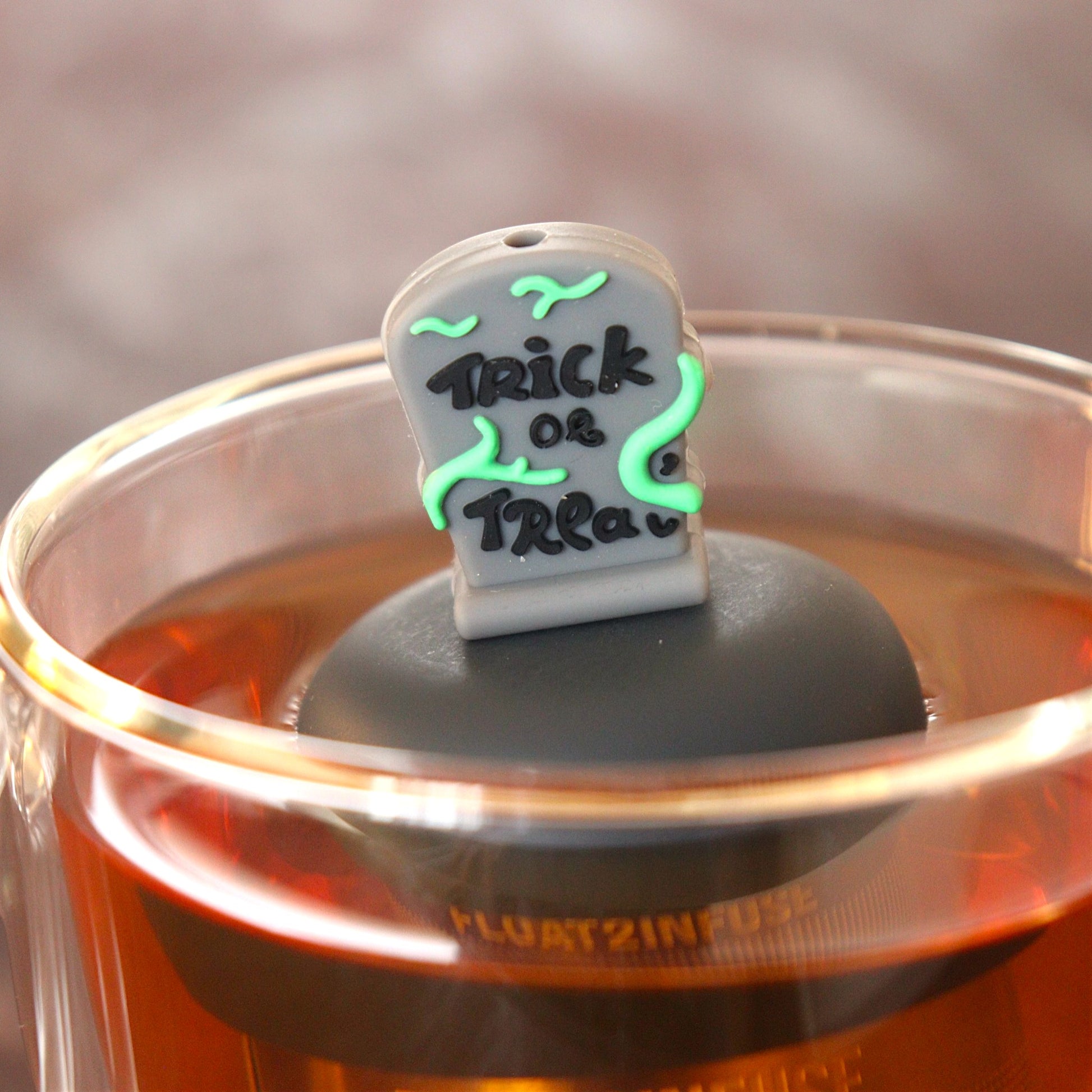 green vines wrap around a halloween themed tombstone with text that reads "trick or treat" tea infuser for loose leaf tea  with a fine mesh stainless steel tea infuser that reads FLOAT2INFUSE on a black base floating in a glass tea mug