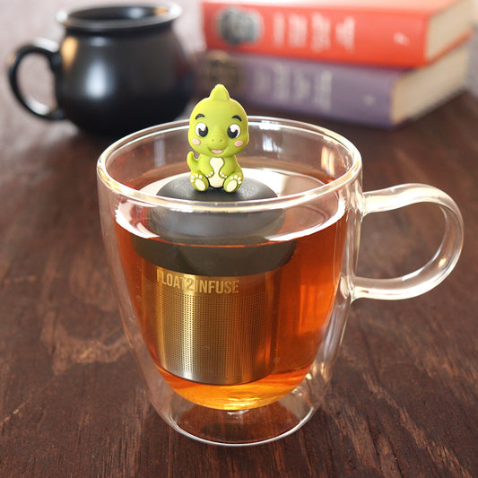 cute baby green dinosaur animal tea infuser for loose leaf tea with a fine mesh stainless steel tea infuser that reads FLOAT2INFUSE on a black base  floating in a glass tea mug with a black cauldron mug and books in the background