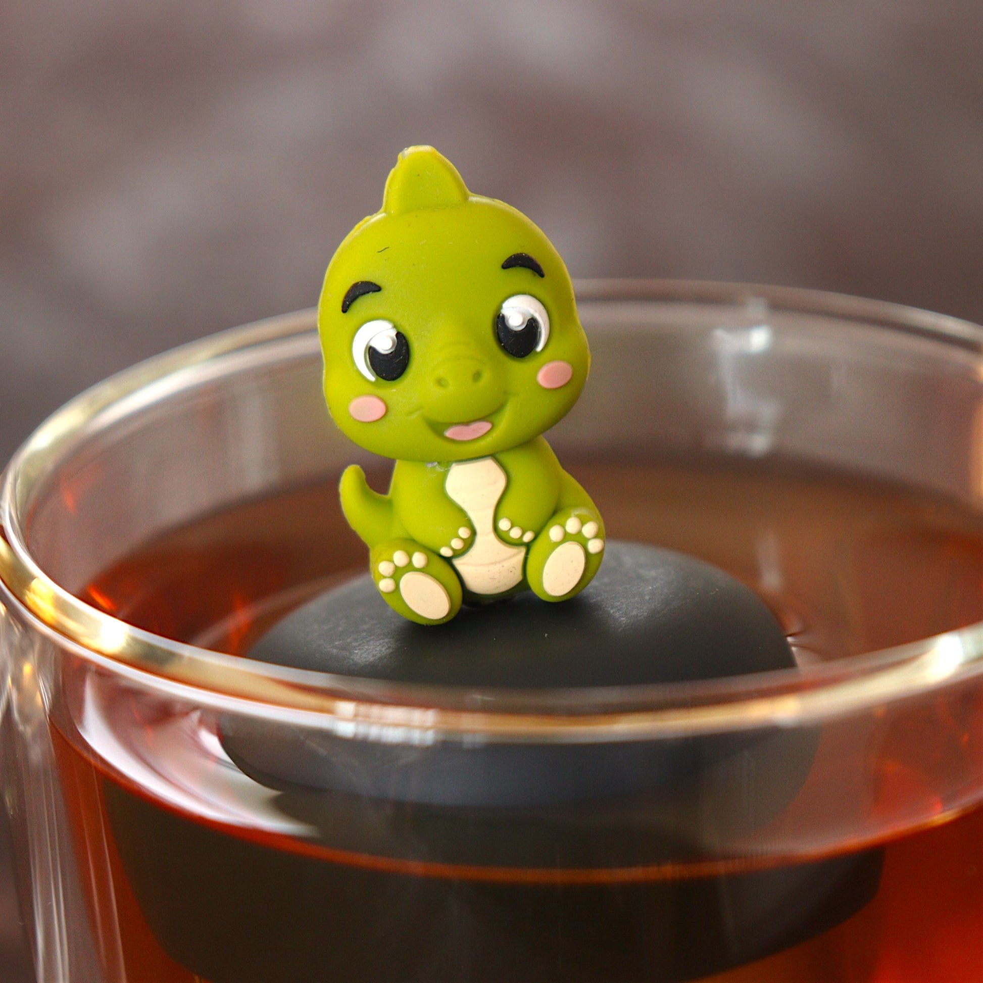 cute baby green dinosaur animal tea infuser for loose leaf tea  with a fine mesh stainless steel tea infuser that reads FLOAT2INFUSE on a black base floating in a glass tea mug
