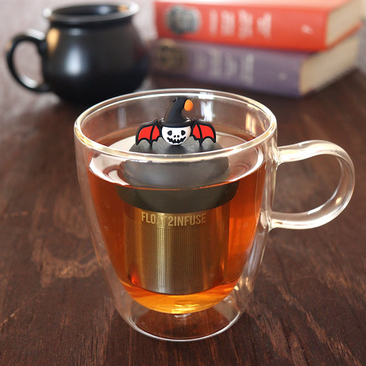 skull with bat wings and halloween hat tea infuser for loose leaf tea with a fine mesh stainless steel tea infuser that reads FLOAT2INFUSE on a black base floating in a glass tea mug with a black cauldron mug and books in the background