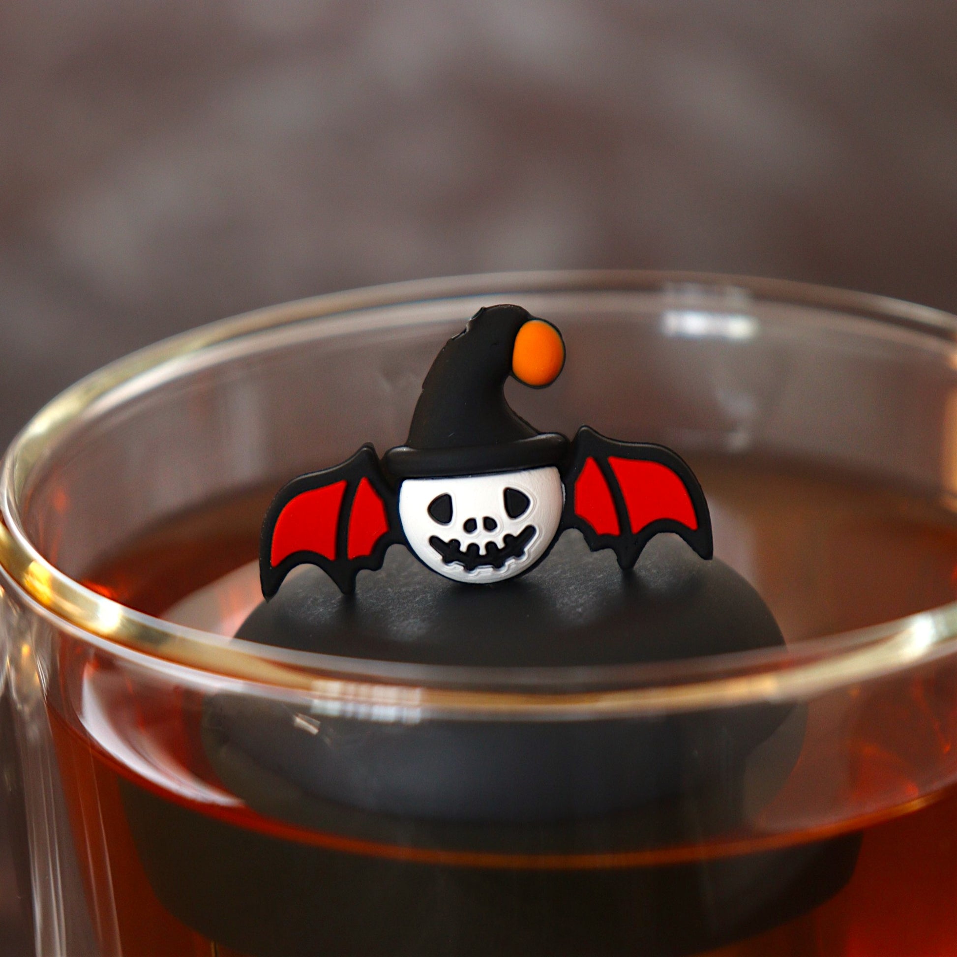 skull with bat wings and halloween hat tea infuser for loose leaf tea  with a fine mesh stainless steel tea infuser that reads FLOAT2INFUSE on a black base floating in a glass tea mug