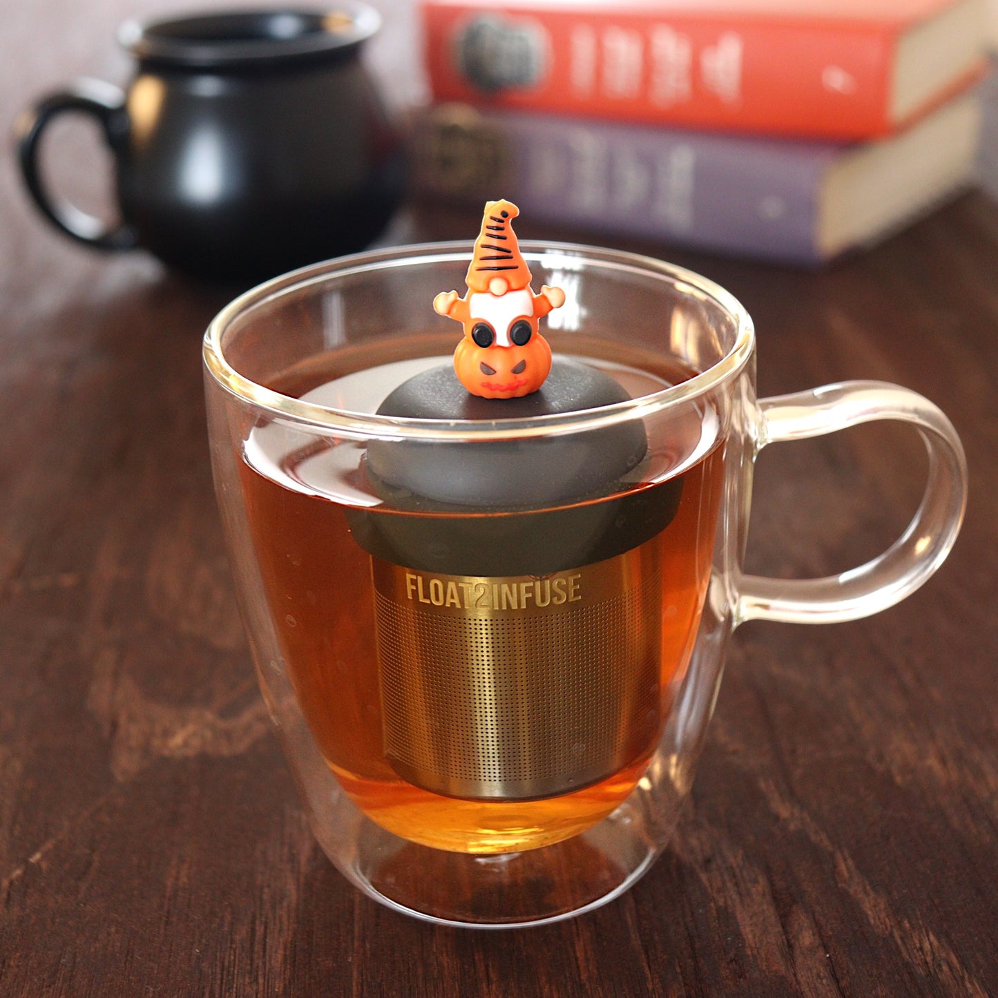 spooky orange gnome on a pumpkin tea infuser for loose leaf tea  with a fine mesh stainless steel tea infuser that reads FLOAT2INFUSE on a black base floating in a glass tea mug with a black cauldron mug and books in the background