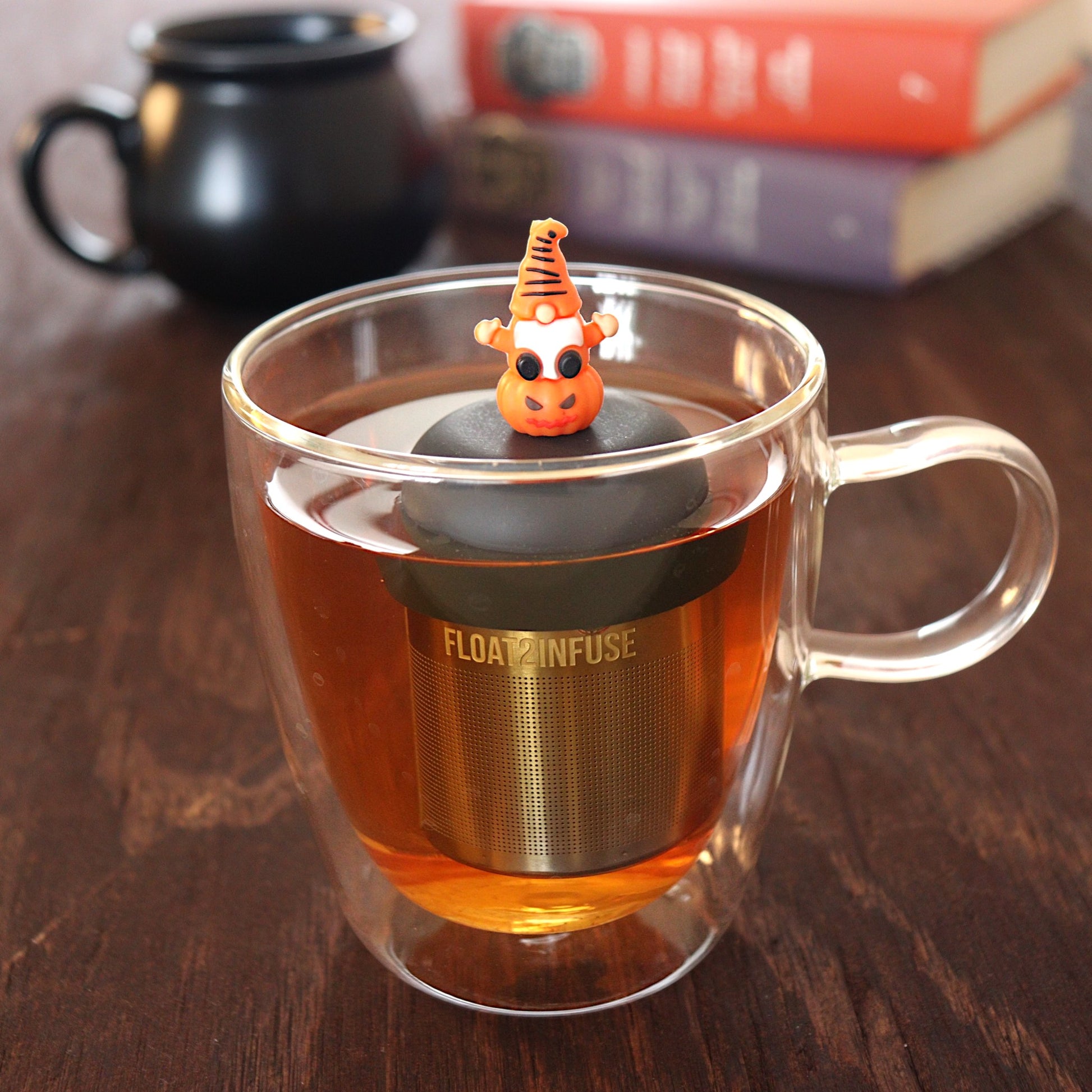 spooky orange gnome on a pumpkin tea infuser for loose leaf tea  with a fine mesh stainless steel tea infuser that reads FLOAT2INFUSE on a black base floating in a glass tea mug with a black cauldron mug and books in the background
