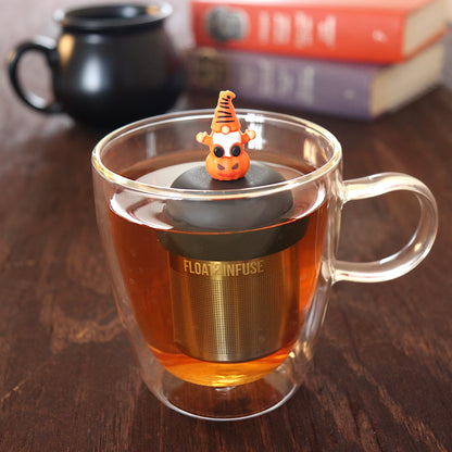 spooky orange gnome on a pumpkin tea infuser for loose leaf tea  with a fine mesh stainless steel tea infuser that reads FLOAT2INFUSE on a black base floating in a glass tea mug with a black cauldron mug and books in the background