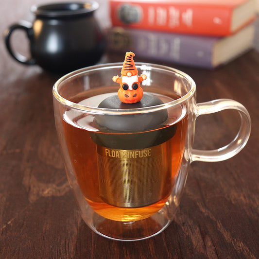 spooky orange gnome on a pumpkin tea infuser for loose leaf tea  with a fine mesh stainless steel tea infuser that reads FLOAT2INFUSE on a black base floating in a glass tea mug with a black cauldron mug and books in the background