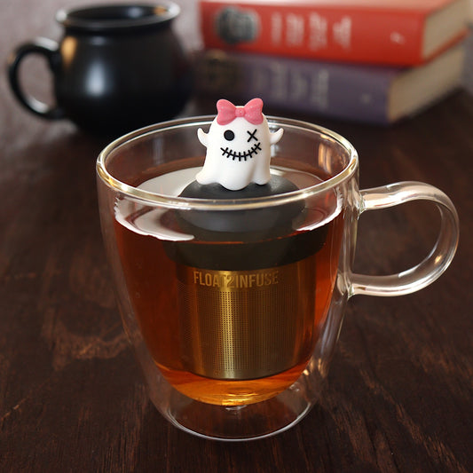 white zombie ghost with a glittery pink bow tea infuser for loose leaf tea  with a fine mesh stainless steel tea infuser that reads FLOAT2INFUSE on a black base floating in a glass tea mug with a black cauldron mug and books in the background