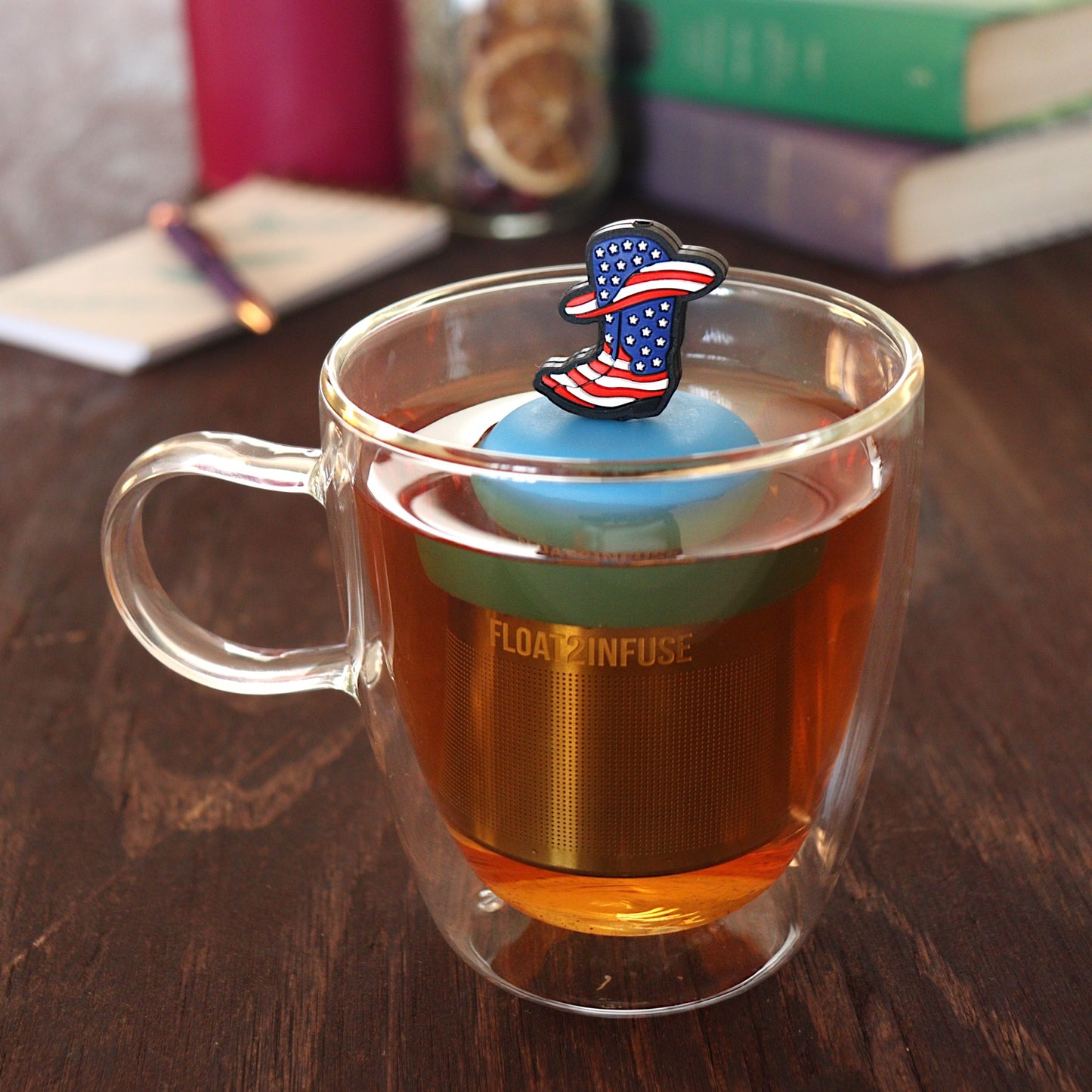 american flag patterned cowboy boots and hat tea infuser for loose leaf tea  with a fine mesh stainless steel tea infuser that reads FLOAT2INFUSE on a blue base floating in a glass tea mug with pen on notebook and loose leaf tea and books in the background
