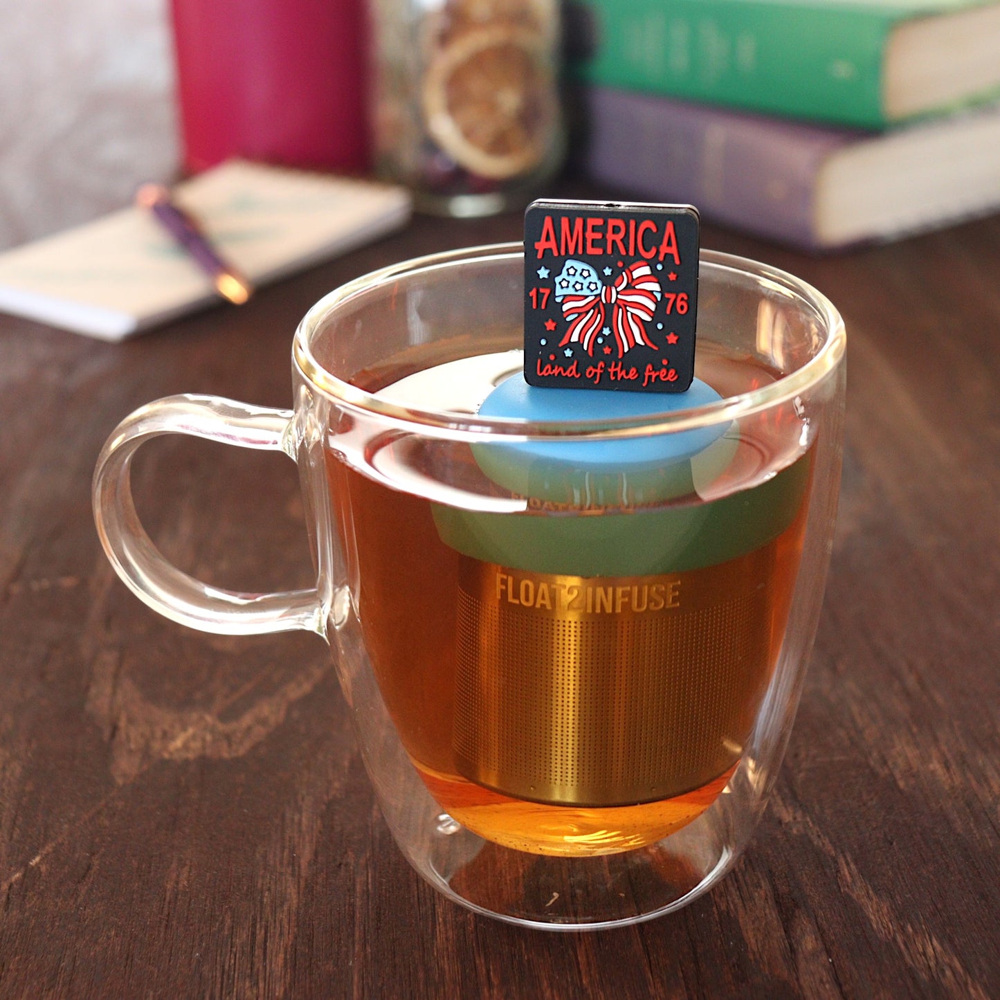 black square with text that reads "america land of the free 1776" that surrounds an american flag bow tea infuser for loose leaf tea  with a fine mesh stainless steel tea infuser that reads FLOAT2INFUSE on a blue base floating in a glass tea mug with pen on notebook and loose leaf tea and books in the background