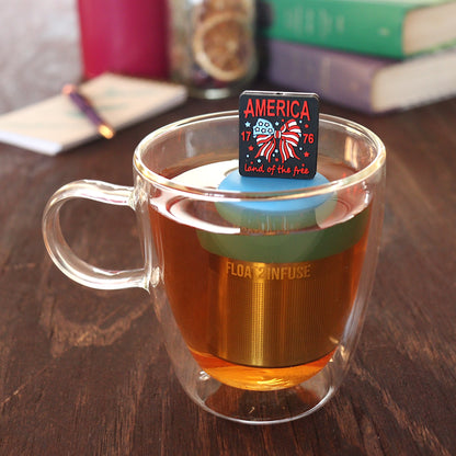 black square with text that reads "america land of the free 1776" that surrounds an american flag bow tea infuser for loose leaf tea  with a fine mesh stainless steel tea infuser that reads FLOAT2INFUSE on a blue base floating in a glass tea mug with pen on notebook and loose leaf tea and books in the background