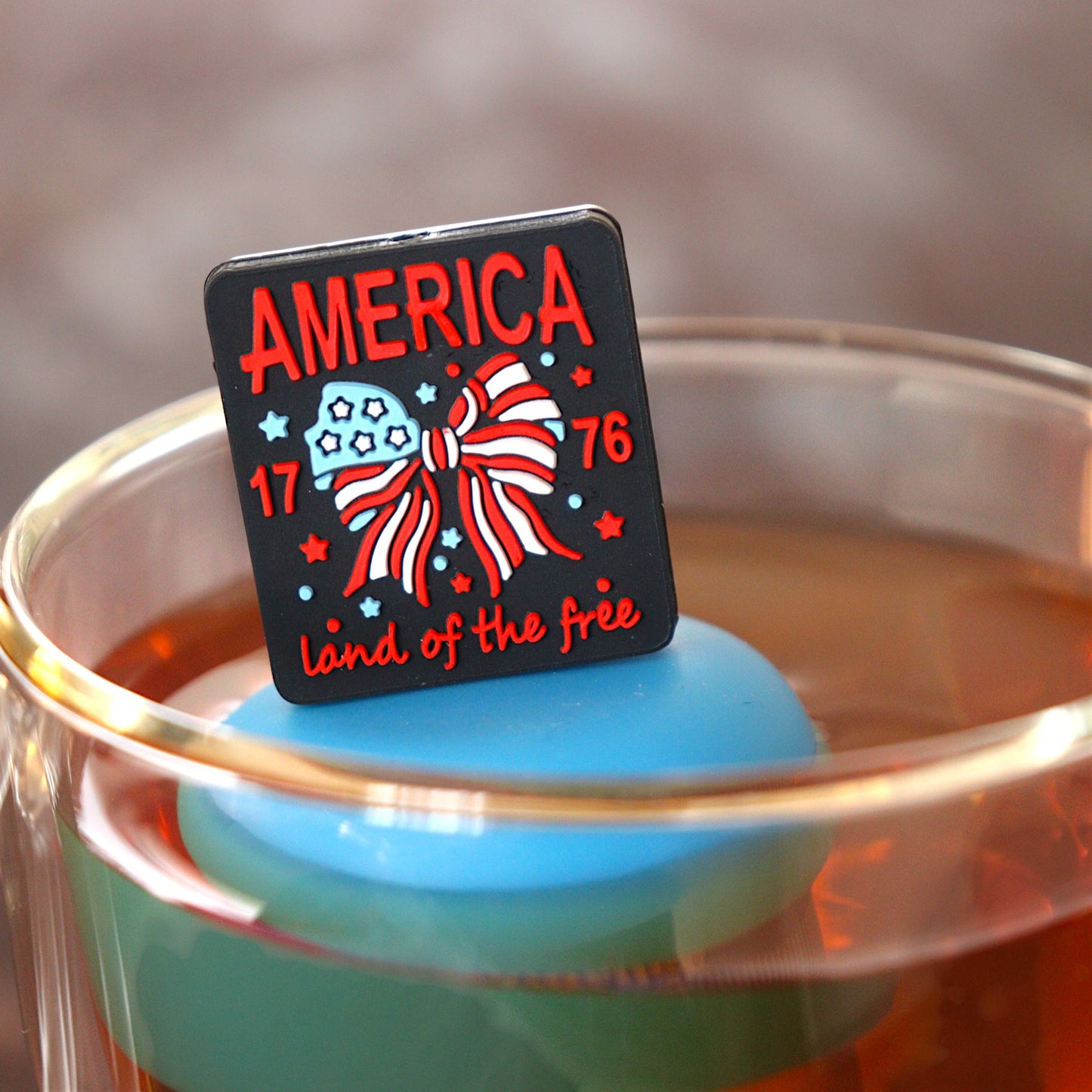 black square with text that reads "america land of the free 1776" that surrounds an american flag bow tea infuser for loose leaf tea  with a fine mesh stainless steel tea infuser that reads FLOAT2INFUSE on a blue base floating in a glass tea mug