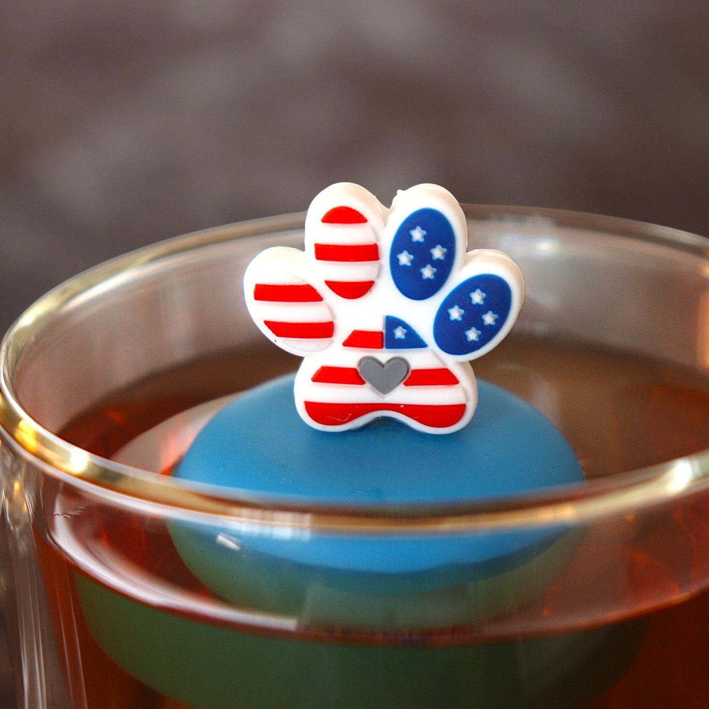 Pawprint with an American flag pattern and grey heart in the center tea infuser for loose leaf tea with a fine mesh stainless steel tea strainer 
that reads FLOAT2INFUSE on a blue base floating in a glass tea mug