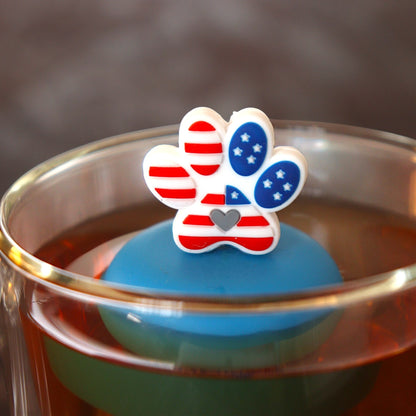 Pawprint with an American flag pattern and grey heart in the center tea infuser for loose leaf tea with a fine mesh stainless steel tea strainer 
that reads FLOAT2INFUSE on a blue base floating in a glass tea mug