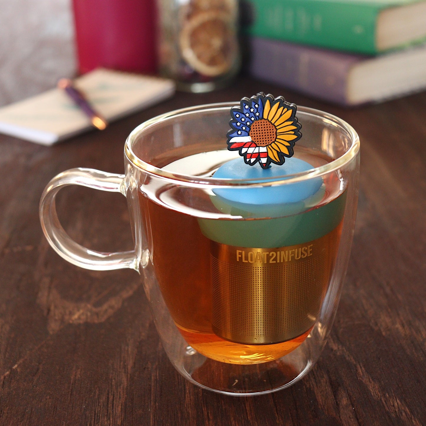 american flag patterned sunflower tea infuser for loose leaf tea  with a fine mesh stainless steel tea infuser that reads FLOAT2INFUSE on a blue base floating in a glass tea mug with pen on notebook and loose leaf tea and books in the background