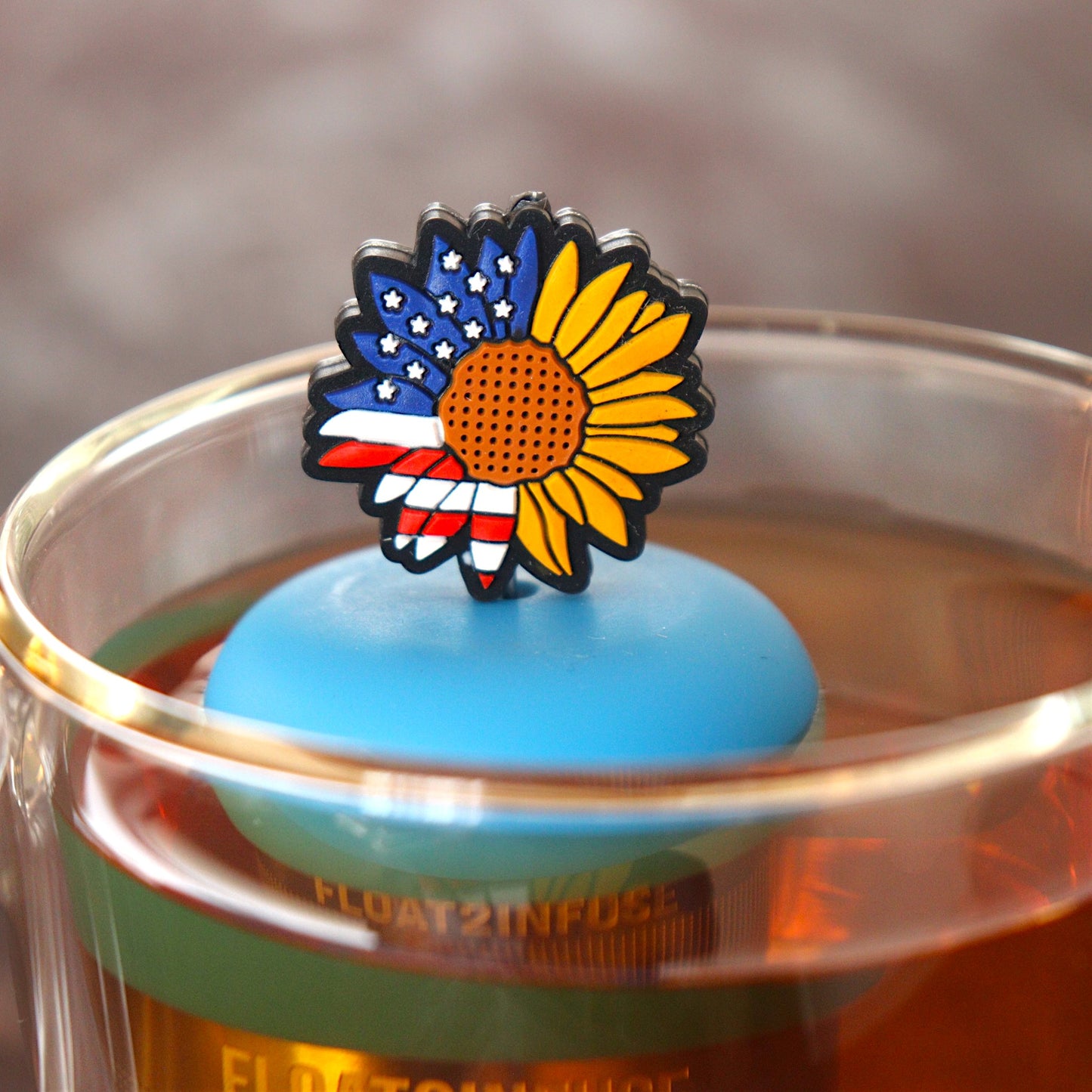 american flag patterned sunflower tea infuser for loose leaf tea with a fine mesh stainless steel tea infuser that reads FLOAT2INFUSE  on a blue base floating in a glass tea mug
