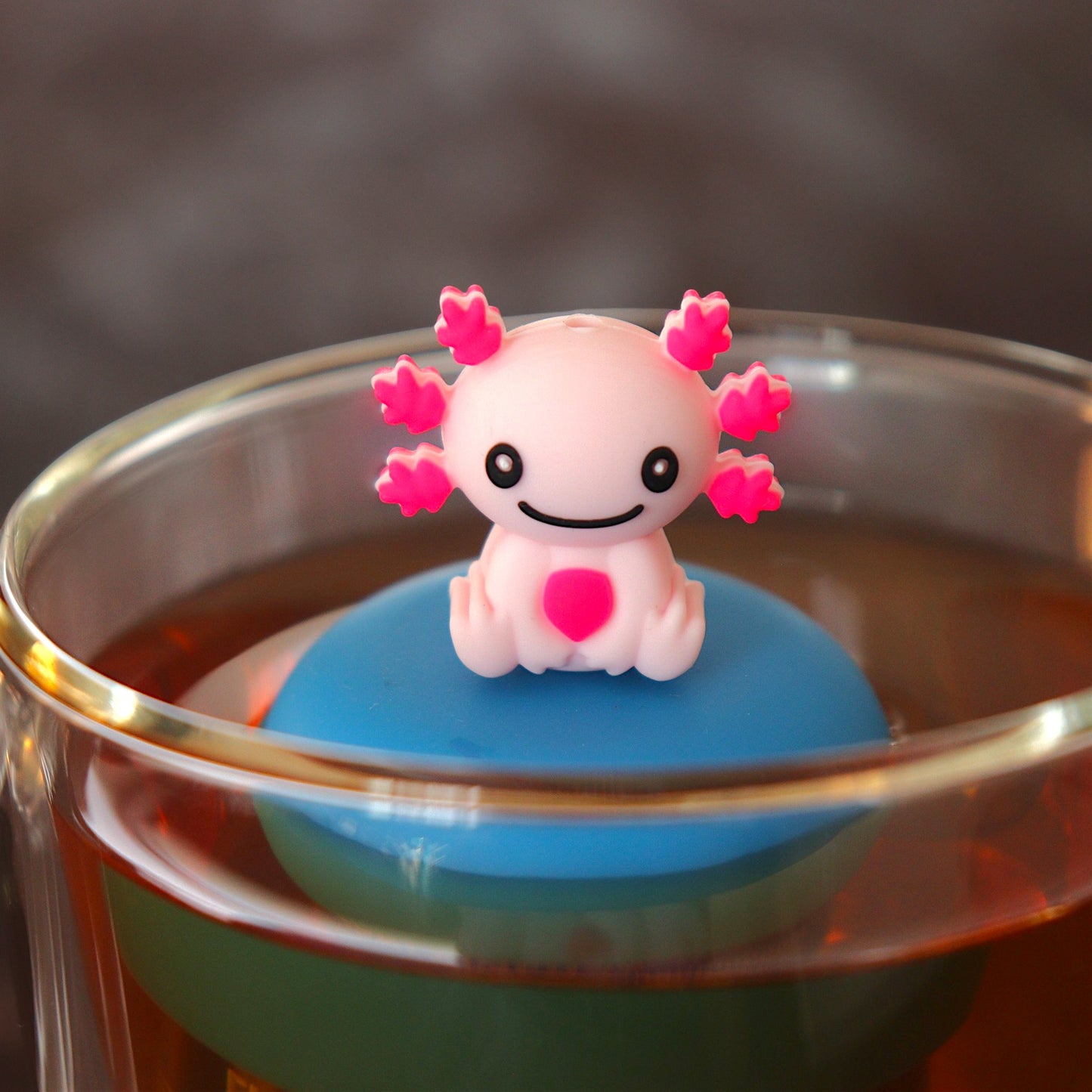 happy pink axolotl tea infuser for loose leaf tea with a fine mesh stainless steel tea strainer that reads FLOAT2INFUSE on a blue base floating in a glass tea mug