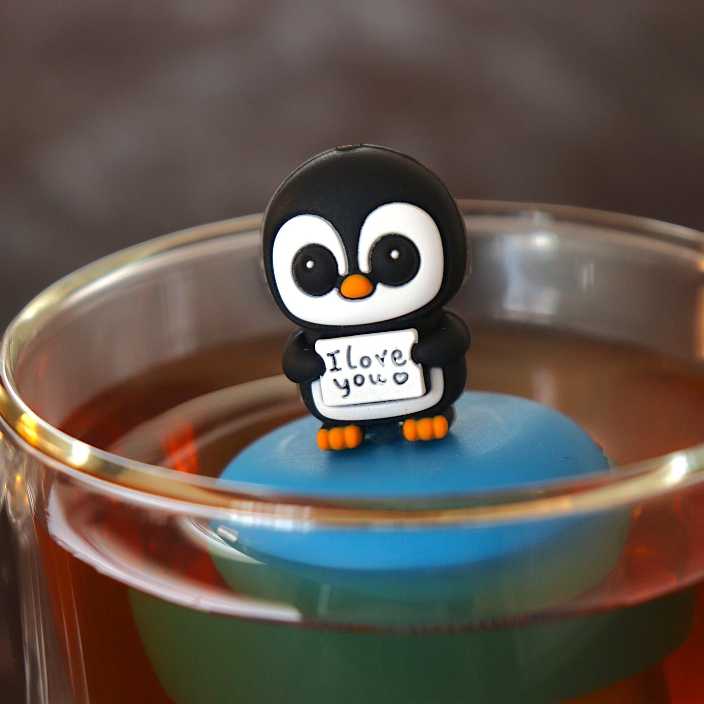 black and white penguin animal holding a sign with text that reads "i love you" tea infuser for loose leaf tea  with a fine mesh stainless steel tea infuser that reads FLOAT2INFUSE on a blue base floating in a glass tea mug