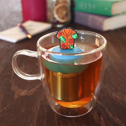 orange hawaiian shirt tea infuser for loose leaf tea on a blue base with a fine mesh stainless steel tea infuser that reads FLOAT2INFUSE floating in a glass tea mug with pen on notebook and loose leaf tea and books in the background