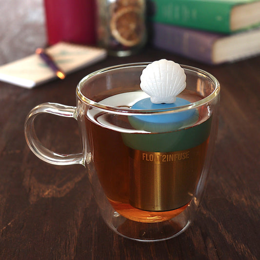 white scallop shell tea infuser for loose leaf tea on a blue base with a fine mesh stainless steel tea infuser that reads FLOAT2INFUSE floating in a glass tea mug with pen on notebook and loose leaf tea and books in the background