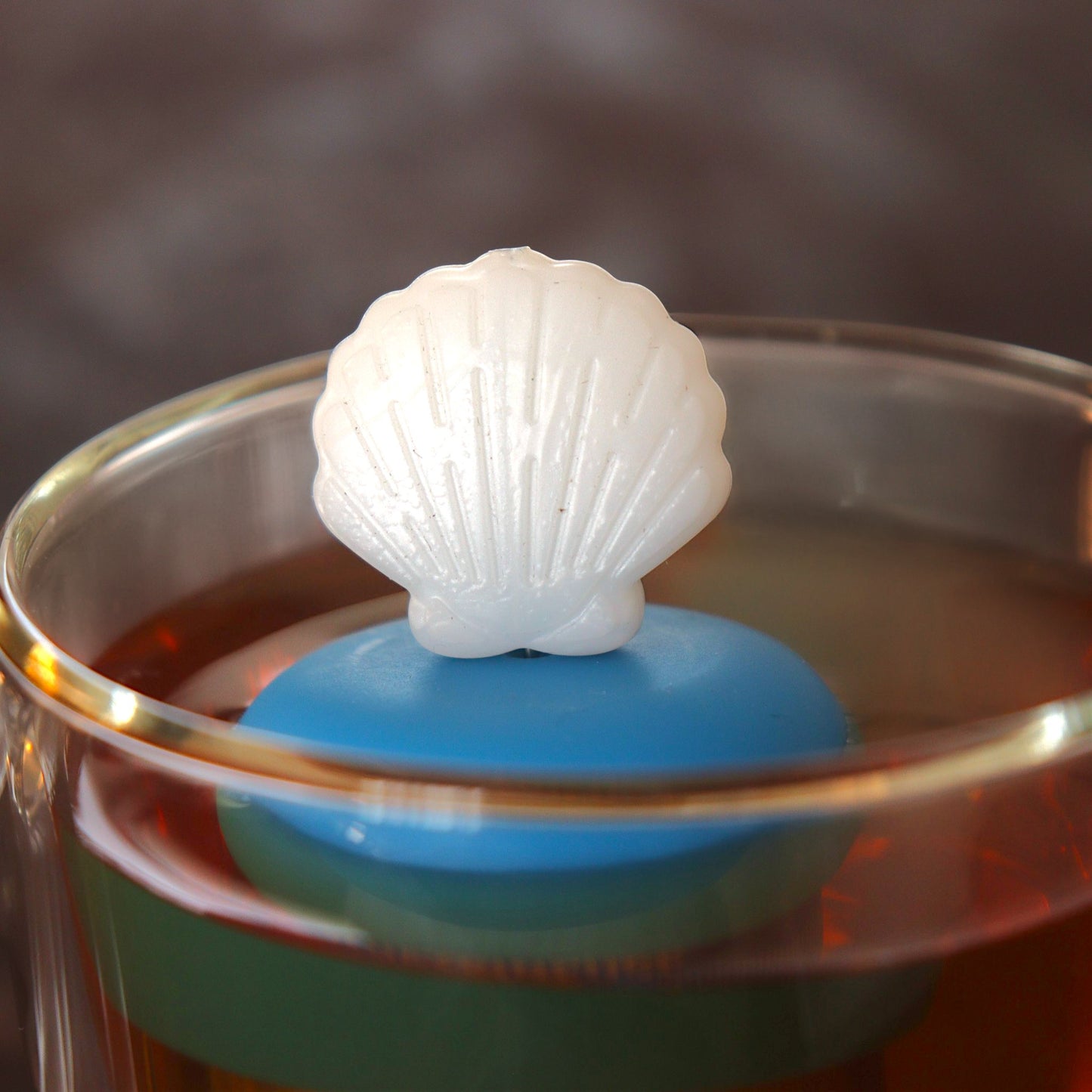 white scallop shell tea infuser for loose leaf tea on a blue base with a fine mesh stainless steel tea infuser that reads FLOAT2INFUSE floating in a glass tea mug