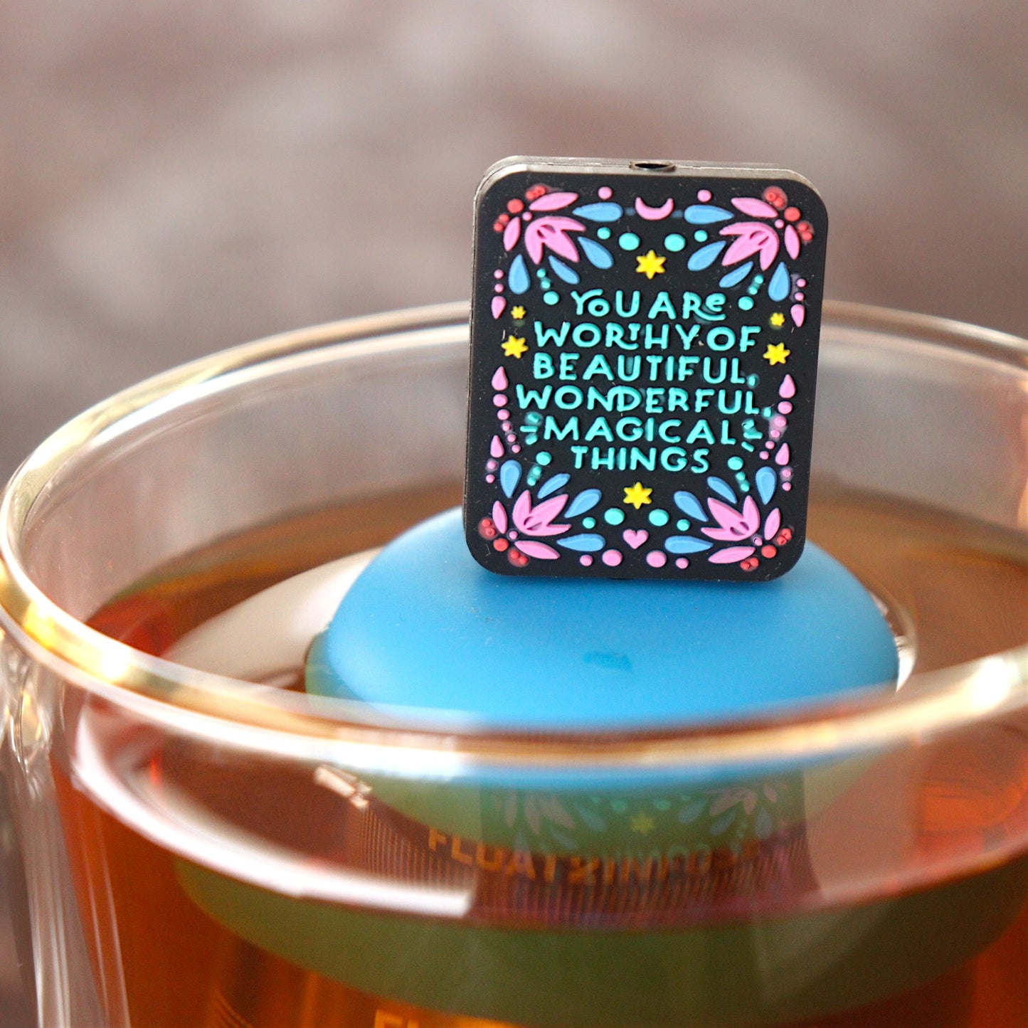 black rectangle with text that reads "you are worthy of beautiful wonderful magical things" tea infuser for loose leaf tea with a fine mesh stainless steel tea infuser that reads FLOAT2INFUSE on a blue base  floating in a glass tea mug
