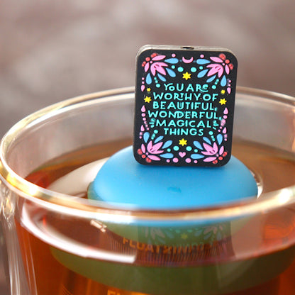 black rectangle with text that reads "you are worthy of beautiful wonderful magical things" tea infuser for loose leaf tea with a fine mesh stainless steel tea infuser that reads FLOAT2INFUSE on a blue base  floating in a glass tea mug