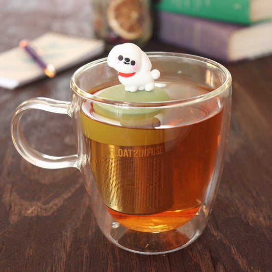 cute white maltese dog tea infuser for loose leaf tea  with a fine mesh stainless steel tea infuser that reads FLOAT2INFUSE on a green base floating in a glass tea mug with pen on notebook and loose leaf tea and books in the background