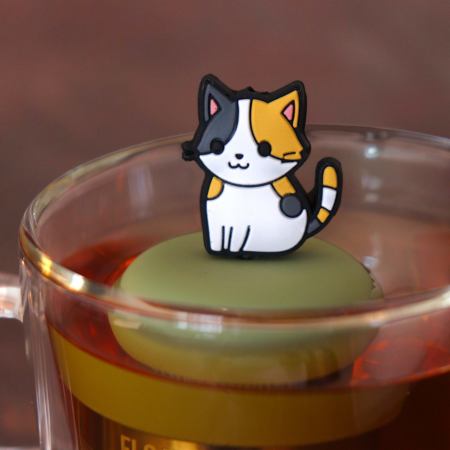 anime Calico cat tea infuser for loose leaf tea  with a fine mesh stainless steel tea infuser that reads FLOAT2INFUSE on a green base floating in a glass tea mug