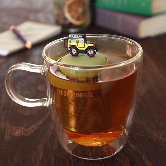 yellow jeep car tea infuser for loose leaf tea  with a fine mesh stainless steel tea infuser that reads FLOAT2INFUSE on a green base floating in a glass tea mug with pen on notebook and loose leaf tea and books in the background
