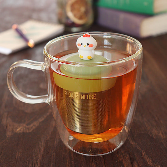 white baby chicken tea infuser for loose leaf tea  with a fine mesh stainless steel tea infuser that reads FLOAT2INFUSE on a green base floating in a glass tea mug with pen on notebook and loose leaf tea and books in the background