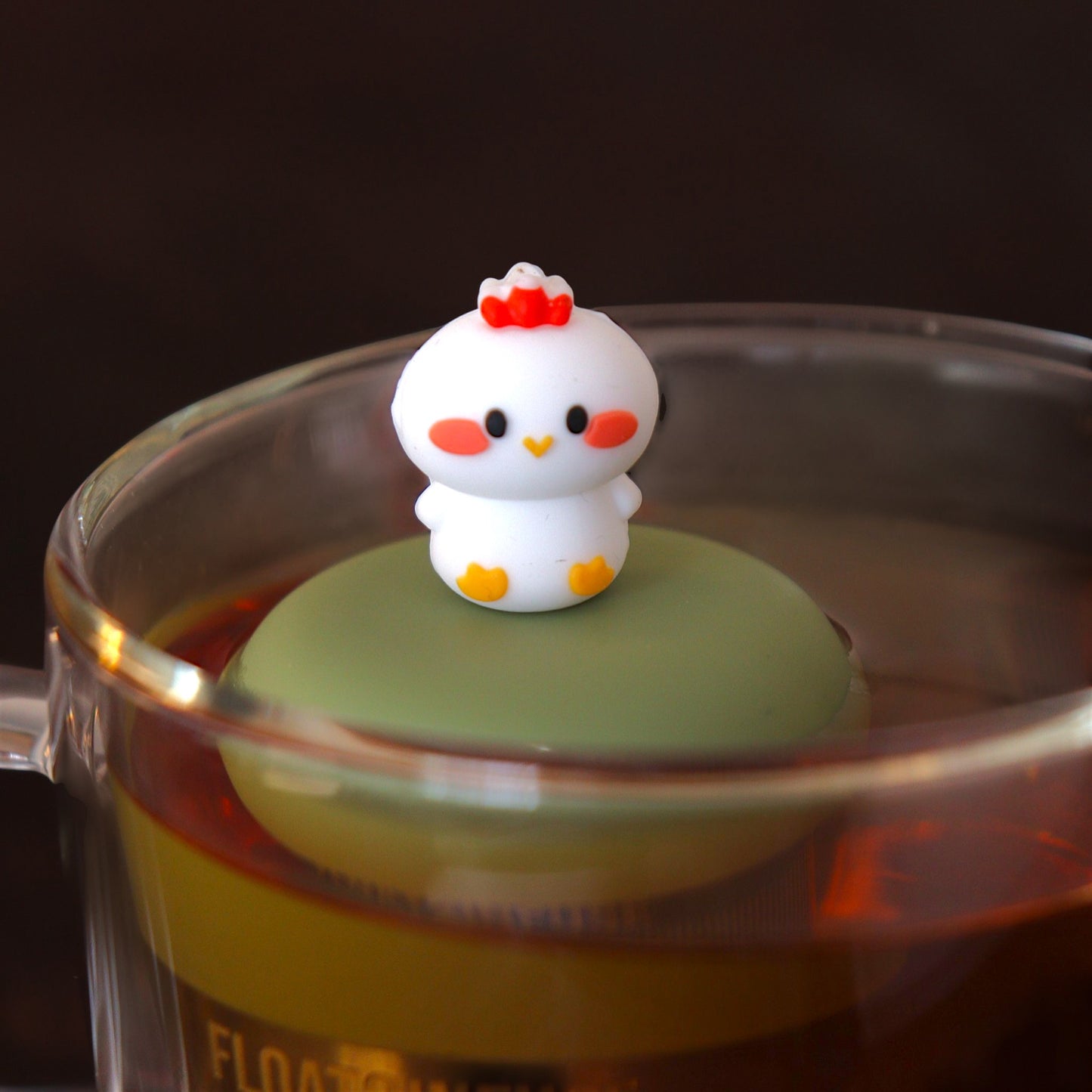 white baby chicken tea infuser for loose leaf tea with a fine mesh stainless steel tea infuser that reads FLOAT2INFUSE on a green base  floating in a glass tea mug