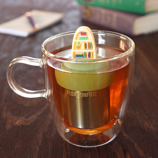blonde plant bookshelf tea infuser for loose leaf tea with a fine mesh stainless steel tea infuser that reads FLOAT2INFUSE on a green base floating in a glass tea mug with pen on notebook and loose leaf tea and books in the background