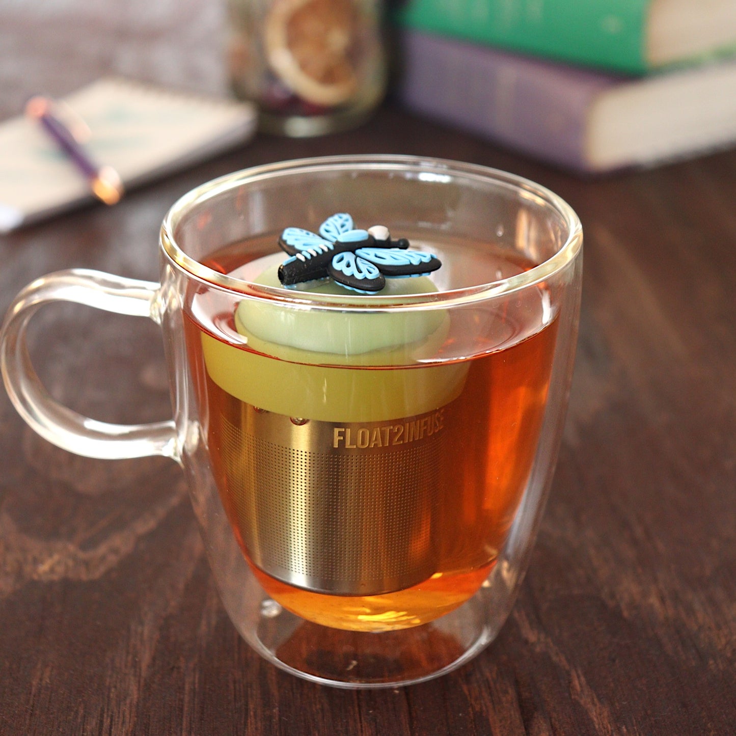 Blue dragonfly tea infuser for loose leaf tea with a fine mesh stainless steel tea strainer 
that reads FLOAT2INFUSE on a green base floating in a glass tea mug 
with pen and notebook and books in the background
