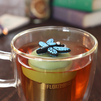 Side view of a blue dragonfly tea infuser for loose leaf tea with a fine mesh stainless steel tea strainer 
that reads FLOAT2INFUSE on a green base floating in a glass tea mug  with books in the background
