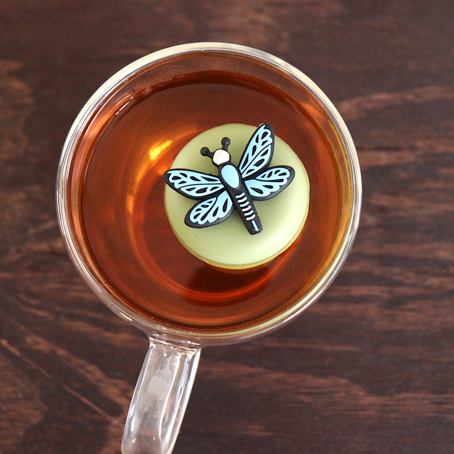 Top view of a blue dragonfly tea infuser for loose leaf tea with a fine mesh stainless steel tea strainer 
that reads FLOAT2INFUSE on a green base floating in a glass tea mug 
