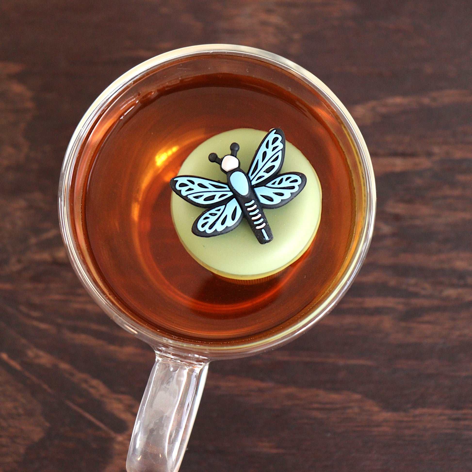 Top view of a blue dragonfly tea infuser for loose leaf tea with a fine mesh stainless steel tea strainer 
that reads FLOAT2INFUSE on a green base floating in a glass tea mug 
