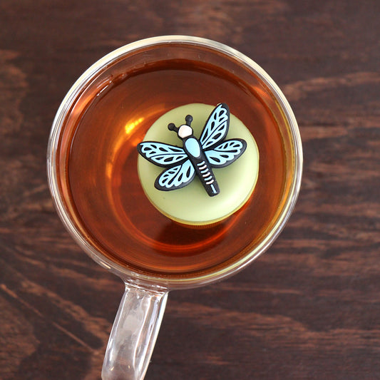 Top view of a blue dragonfly tea infuser for loose leaf tea with a fine mesh stainless steel tea strainer 
that reads FLOAT2INFUSE on a green base floating in a glass tea mug 
