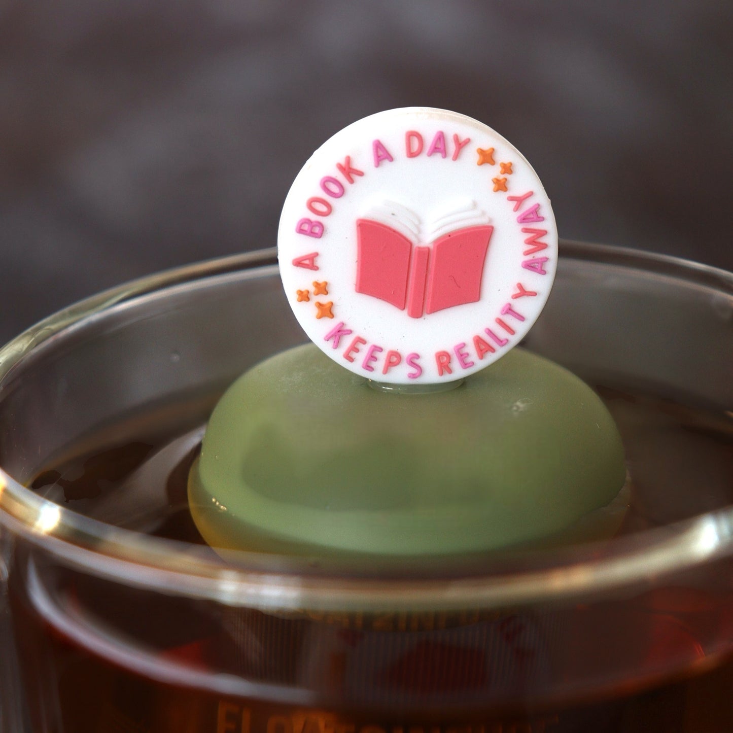 white circle that reads "a book a day keeps the reality away" with a centered pink book tea infuser for loose leaf tea with a fine mesh stainless steel tea infuser that reads FLOAT2INFUSE  on a green base floating in a glass tea mug