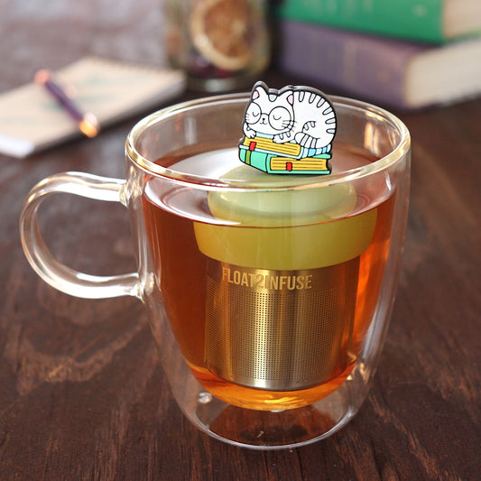 grey cat animal with glasses resting on a stack of green books tea infuser for loose leaf tea on a green base with a fine mesh stainless steel tea infuser that reads FLOAT2INFUSE floating in a glass tea mug with pen on notebook and loose leaf tea and books in the background