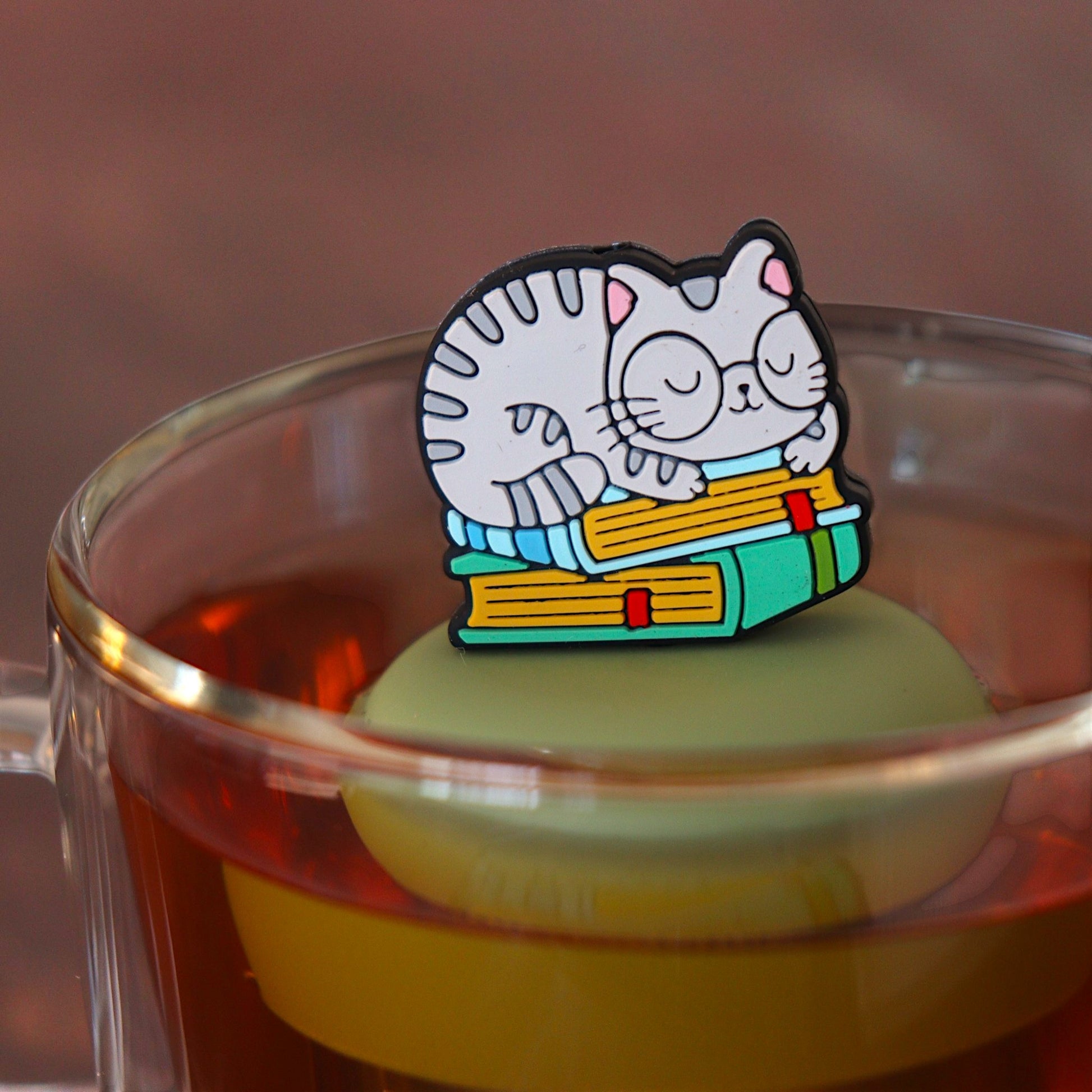 grey cat animal with glasses resting on a stack of green books tea infuser for loose leaf tea with a fine mesh stainless steel tea infuser that reads FLOAT2INFUSE on a green base floating in a glass tea mug 