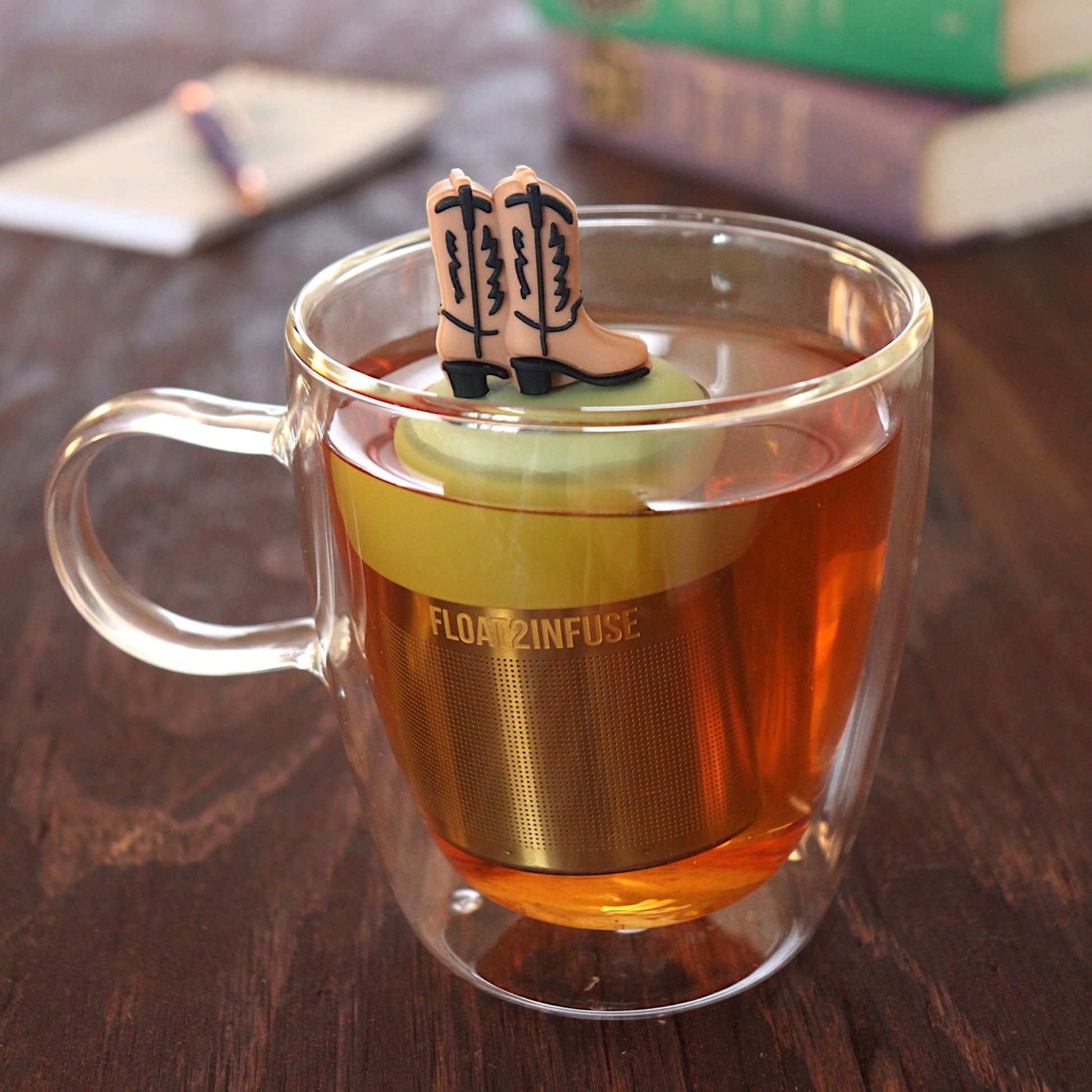 brown cowboy boots tea infuser for loose leaf tea  with a fine mesh stainless steel tea infuser that reads FLOAT2INFUSE on a green base floating in a glass tea mug with pen on notebook and books in the background